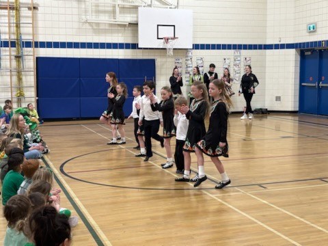 McConnel Irish Dancers perform in a school gymasium There are approximately 12 dancers Some children can be seen seated on the floor watching them.
