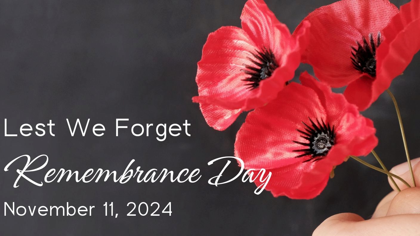 A hand holding three red poppies against a chalkboard background with text that reads “Lest We Forget – Remembrance Day – November 11, 2024”
